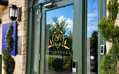 Main entrance to SKYLARANNA Resort & Spa, featuring glass doors with the resort logo, stone exterior, and decorative greenery.