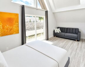 Cabin bedroom and sitting area at SKYLARANNA Resort & Spa, featuring a queen bed, sofa, vaulted ceiling, colourful artwork, and sliding doors providing natural light.