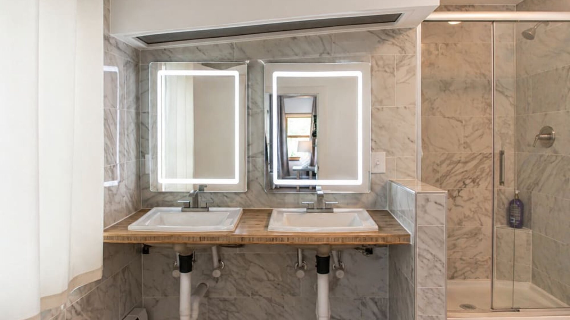 Guest bathroom at SKYLARANNA Resort & Spa, featuring double sinks, illuminated mirrors, marble-style finishes, and a glass-enclosed shower.