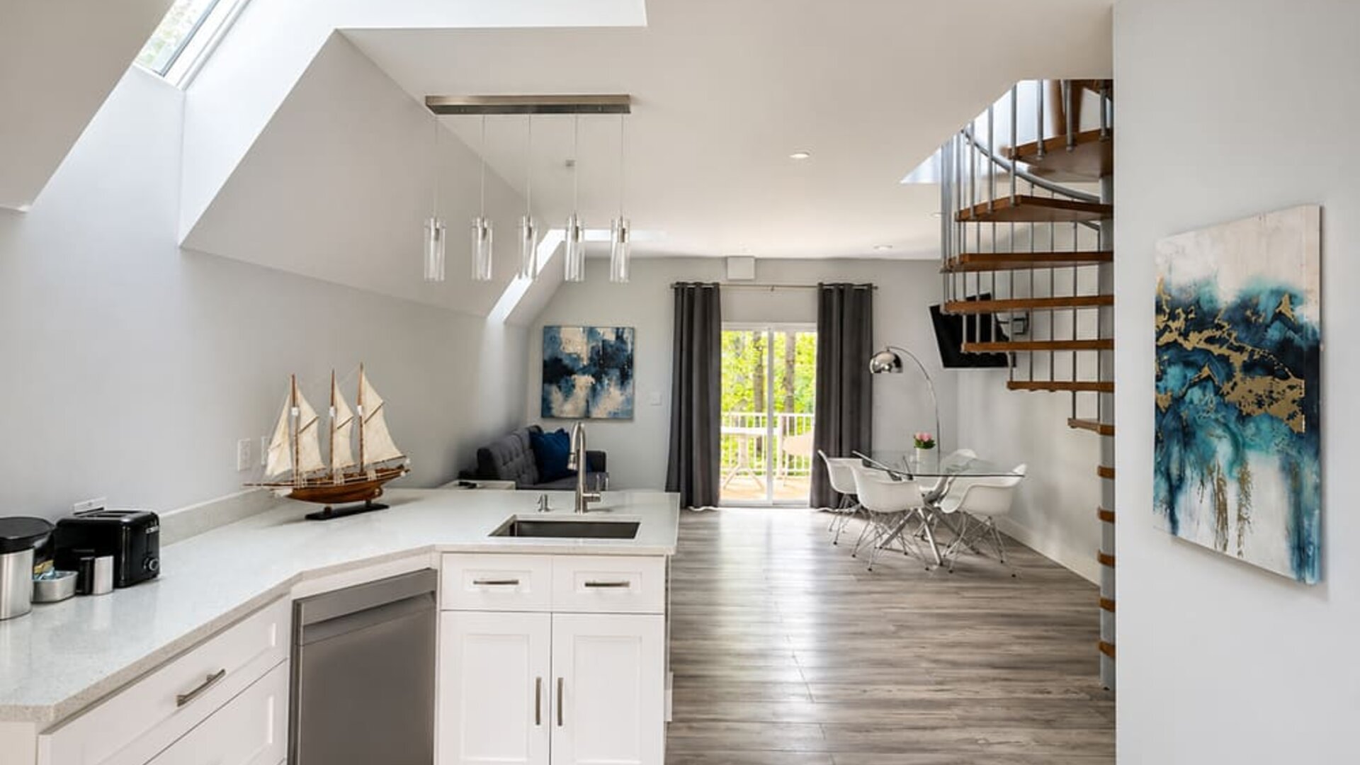 Modern cabin kitchen and open-plan interior at SKYLARANNA Resort & Spa, featuring white cabinetry, an island with sink, dining area, spiral staircase, and contemporary décor.
