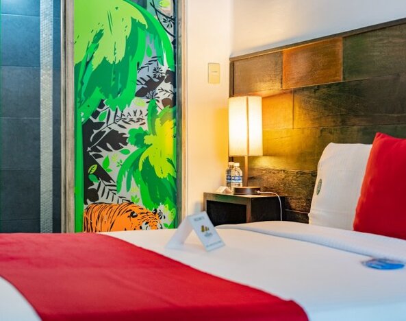 Bright guest room featuring a large bed, exposed brick accent wall, colourful cushions, television, and abundant natural light at Casa Anzures Suites.