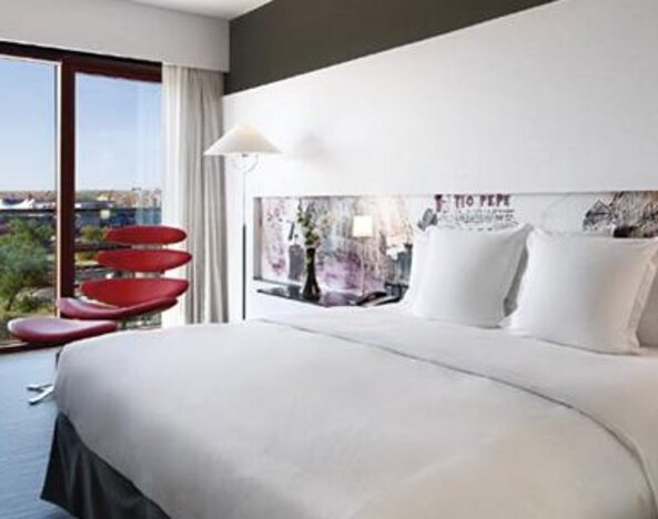 Contemporary guest room featuring a large bed, dedicated workspace, colourful accent seating, and expansive windows with outdoor views at Hilton Madrid Aeropuerto.