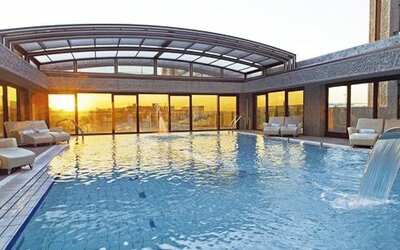Spacious indoor swimming pool featuring a waterfall feature, poolside loungers, and expansive windows with sunset views at Hilton Madrid Aeropuerto.