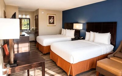 Bright hotel guest room with two white beds against a deep blue accent wall, dark wood headboards, bedside lamp, desk, armchair, and vanity area.