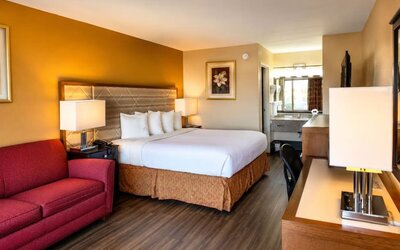 Comfortable hotel guest room with a king bed, white bedding, warm gold accent wall, red sofa, bedside lamps, work desk, wood flooring, and vanity area.
