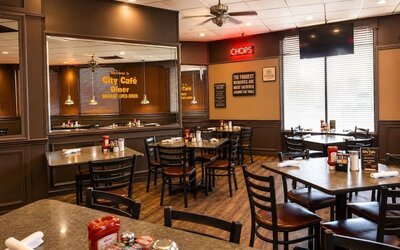 Warmly lit hotel restaurant with dark wood paneling, mirrored walls, multiple dining tables, black chairs, ceiling fans, and large windows with blinds.