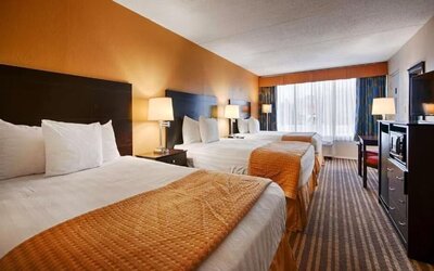 Spacious hotel room with three white beds arranged in a row, orange textured bed runners, bedside lamps, large window with curtains, television, microwave, and storage unit.