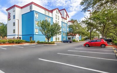 Exterior view of a multi-story hotel with white and blue walls, red roof trim, landscaped grounds, mature trees, and a paved parking lot with several vehicles.