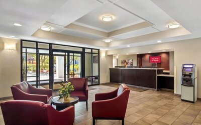 Bright hotel lobby with four red leather armchairs around a small table, tiled flooring, reception desk, ATM, and glass entrance doors overlooking landscaped grounds.