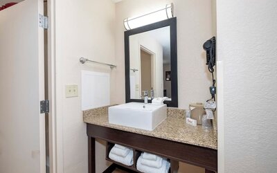 Hotel bathroom vanity with a square white vessel sink, granite countertop, large black-framed mirror, wall-mounted hair dryer, towels, and toiletries.