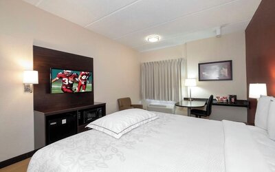 Bright hotel guest room with a king-size bed, white bedding, wall-mounted TV, mini-fridge, microwave, desk, chair, lamps, and curtained window.