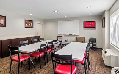 Hotel meeting room with two long conference tables, black chairs with red seats, whiteboard, storage cabinets, office chairs, large window, and wall-mounted air-conditioning units.