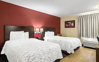 Hotel guest room with two beds dressed in white patterned bedding, dark wood headboards, red accent wall, bedside lamps, curtained window, and air-conditioning unit.