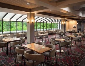Spacious hotel dining area featuring multiple tables, comfortable seating, and expansive windows overlooking lush greenery at Wyndham Garden Pittsburgh Airport.