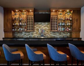 Contemporary hotel bar with a long black counter, blue upholstered bar stools, illuminated wooden shelving stocked with bottles, built-in wine racks, beer taps, and a wall-mounted television.