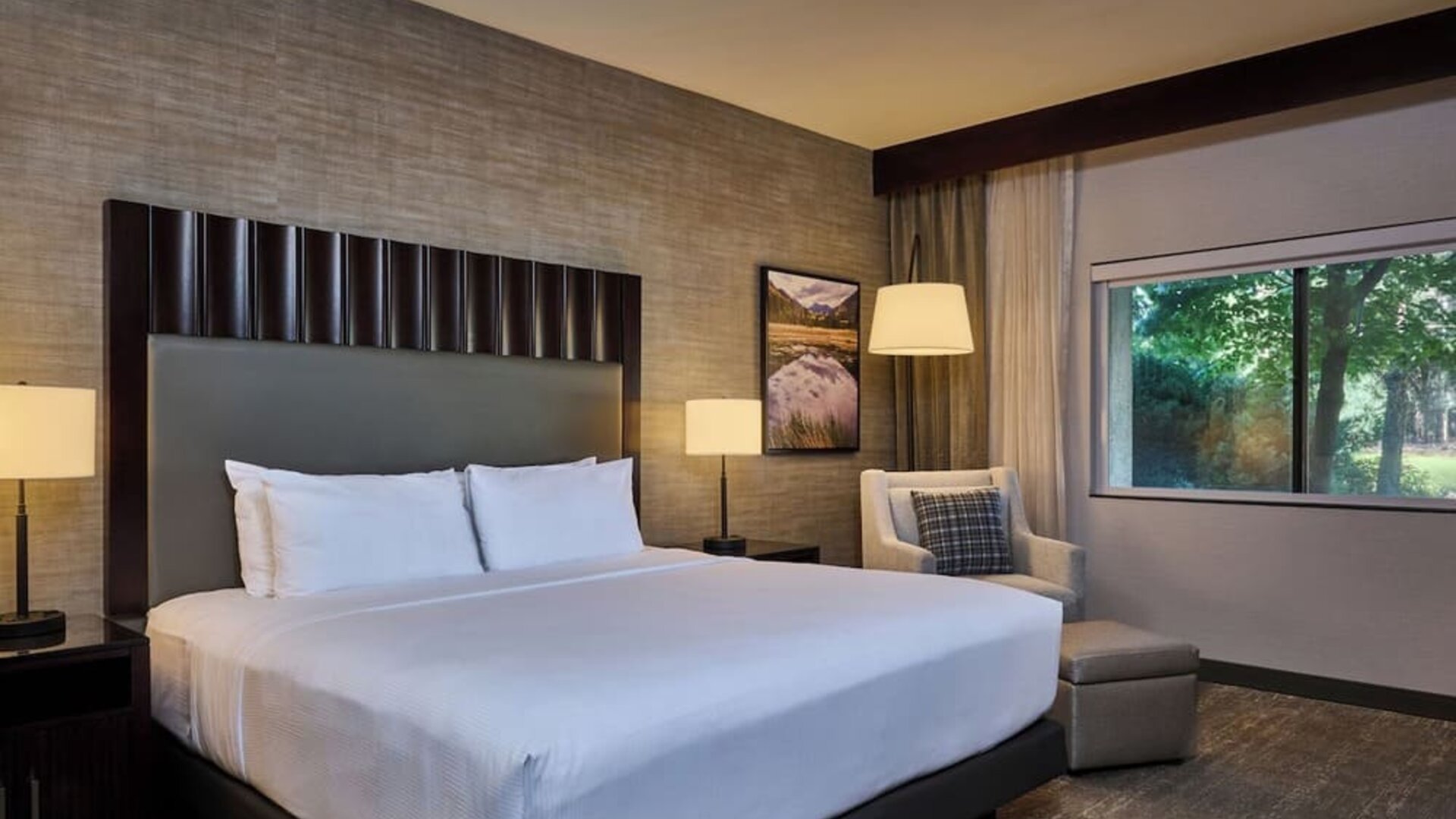 Modern hotel room with a king bed dressed in crisp white linens, tall dark headboard, bedside lamps, upholstered armchair and ottoman, framed artwork, and a large window overlooking green trees.