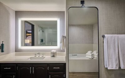 Modern hotel bathroom with a wide illuminated vanity mirror, white countertop and sink, dark wood cabinetry, towel rack, and a bathtub visible through a tall wall mirror.
