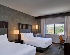 Modern hotel guest room with two beds dressed in white linens, dark upholstered headboards, bedside lighting, a work desk and chair, and a large window overlooking a wooded landscape.