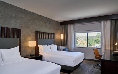 Modern hotel guest room with two beds dressed in white linens, dark upholstered headboards, bedside lighting, a work desk and chair, and a large window overlooking a wooded landscape.
