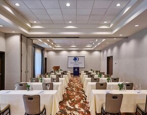Spacious hotel meeting room arranged classroom-style with rows of white-covered tables and chairs, water glasses, small plants, a podium, and a DoubleTree by Hilton presentation screen.