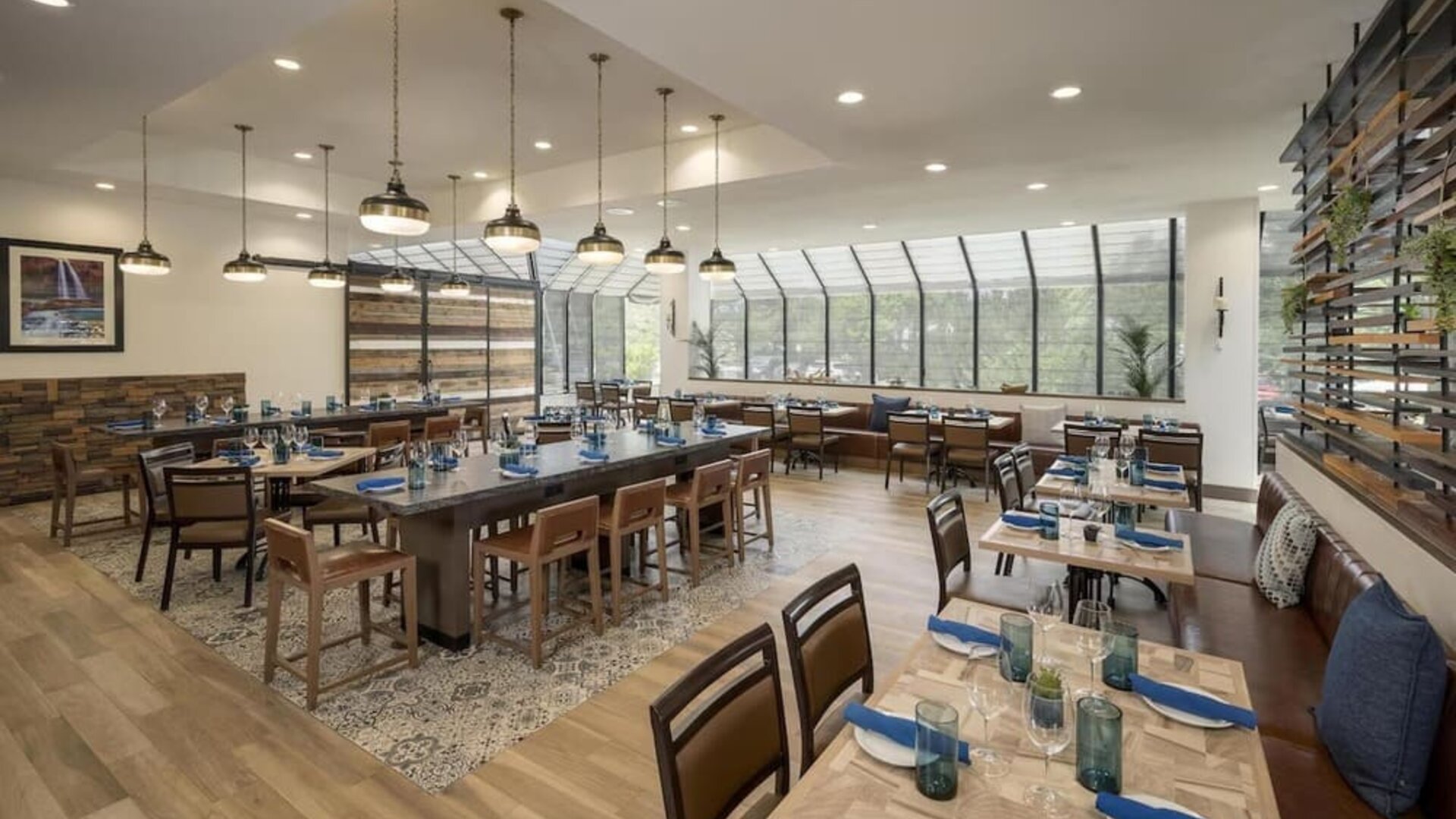 Bright contemporary hotel restaurant with neatly set wooden tables, blue napkins, pendant lighting, patterned flooring, booth seating, and large windows overlooking surrounding greenery.