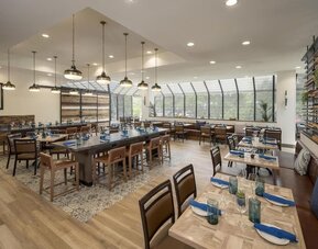 Bright contemporary hotel restaurant with neatly set wooden tables, blue napkins, pendant lighting, patterned flooring, booth seating, and large windows overlooking surrounding greenery.