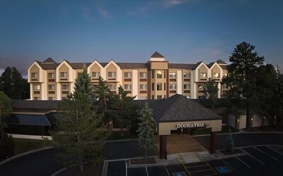 Elevated dusk view of the DoubleTree hotel exterior with illuminated entrance signage, multi-story beige facade, surrounding pine trees, dark parking area, and accessible parking spaces near the entrance.