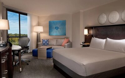 Spacious hotel room with a king bed, white linens, dark upholstered headboard, sofa with colorful accent pillows, work desk and chair, blue artwork, and a large window overlooking trees.