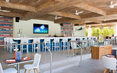 Open-air resort bar with long white counter, blue-and-white stools, beverage taps, colorful coastal decor, dining tables, exposed wood ceiling beams, and multiple ceiling fans.