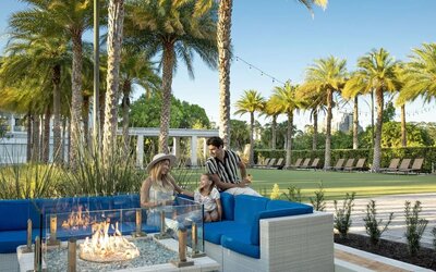 Outdoor resort seating area with bright blue cushioned sofas surrounding a glass-enclosed fire pit, framed by palm trees, landscaped grounds, string lights, and rows of lounge chairs.