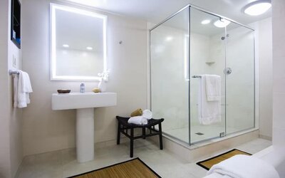 Bright hotel bathroom with illuminated vanity mirror, pedestal sink, glass-enclosed walk-in shower, white towels, bench, neutral tile flooring, and simple contemporary decor.