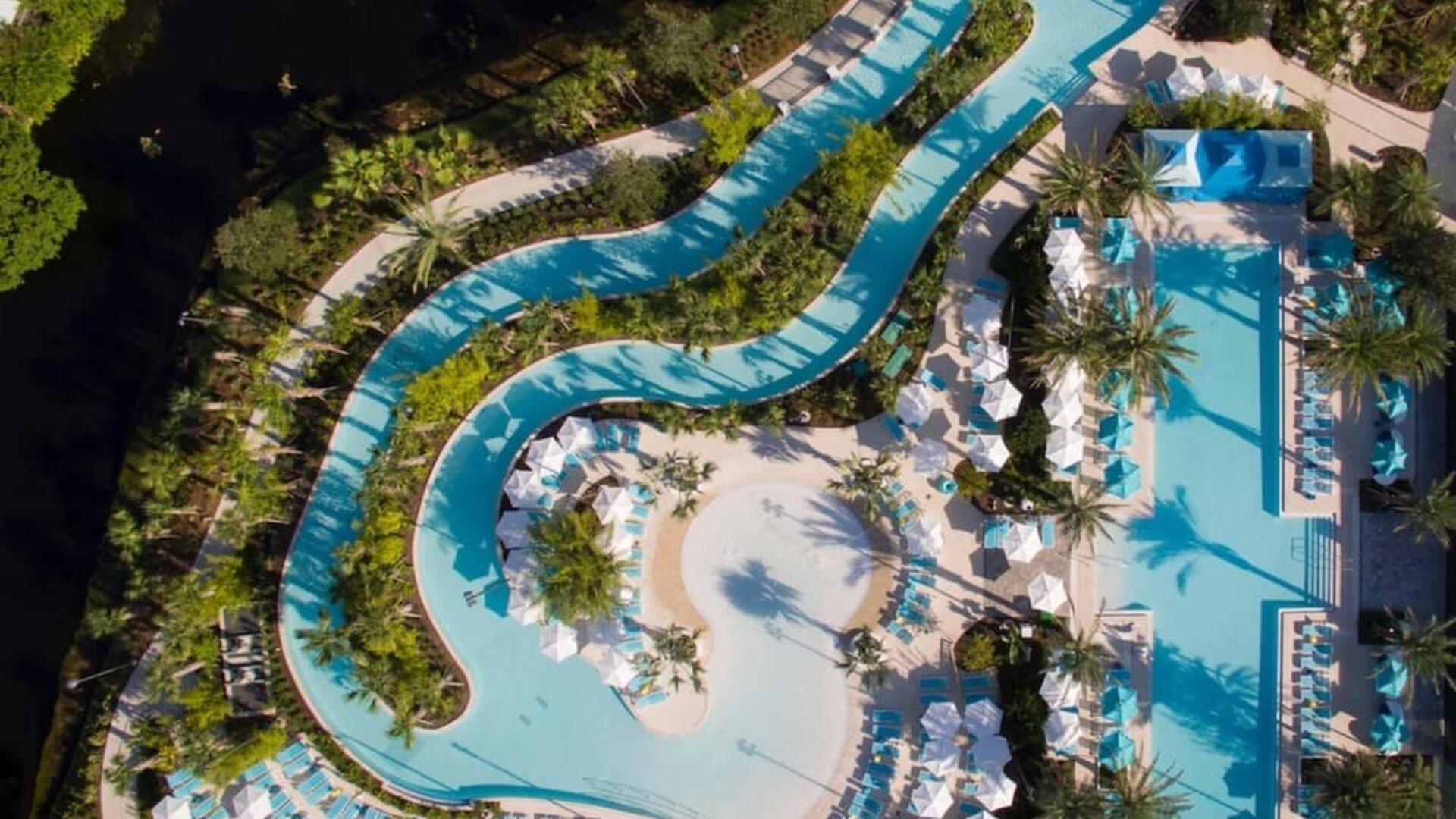Aerial view of an expansive resort pool complex featuring winding lagoon-style pools, a large main pool, tropical landscaping, palm trees, lounge chairs, umbrellas, and cabanas.