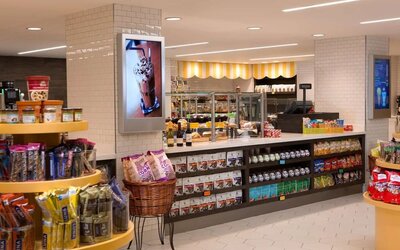 Bright hotel market with shelves and displays stocked with snacks, drinks, packaged foods, and grab-and-go items, plus a service counter and refrigerated cases.
