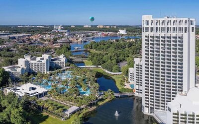 Aerial view of a tall white resort tower beside landscaped grounds, winding waterways, palm-lined swimming pools, and nearby buildings extending across the surrounding area.