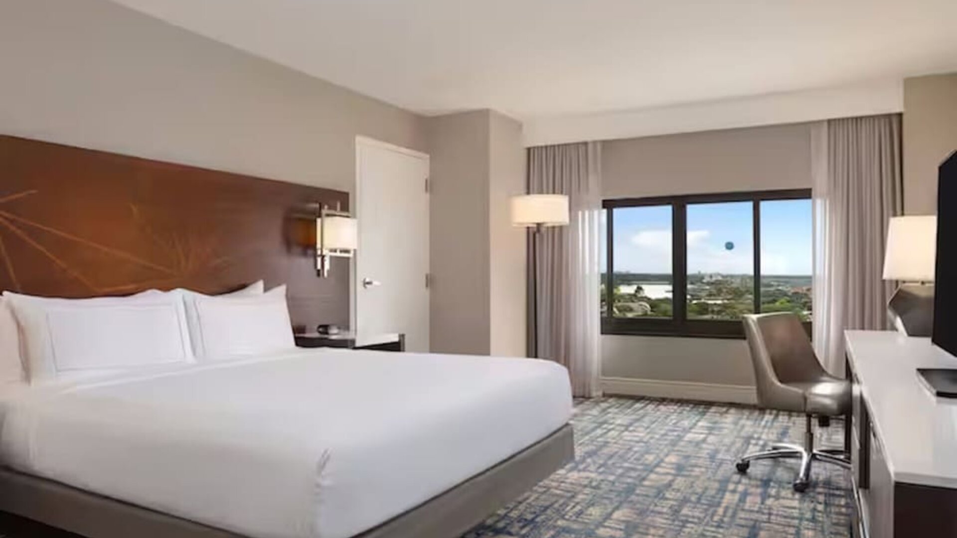 Spacious hotel room with king bed, white linens, large window with an elevated outdoor view, work desk and chair, flat-screen TV, bedside lighting, and patterned carpet.