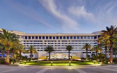 Large multi-story hotel exterior at dusk with illuminated windows and entrance, landscaped circular lawn, mature palm trees, pathways, and exterior lighting.