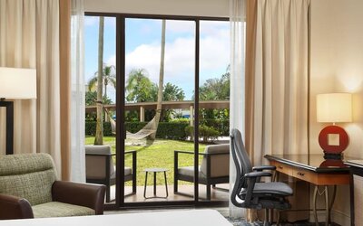 Hotel guest room with desk and ergonomic chair beside sliding glass doors opening to a furnished patio, green lawn, palm trees, and a hammock.