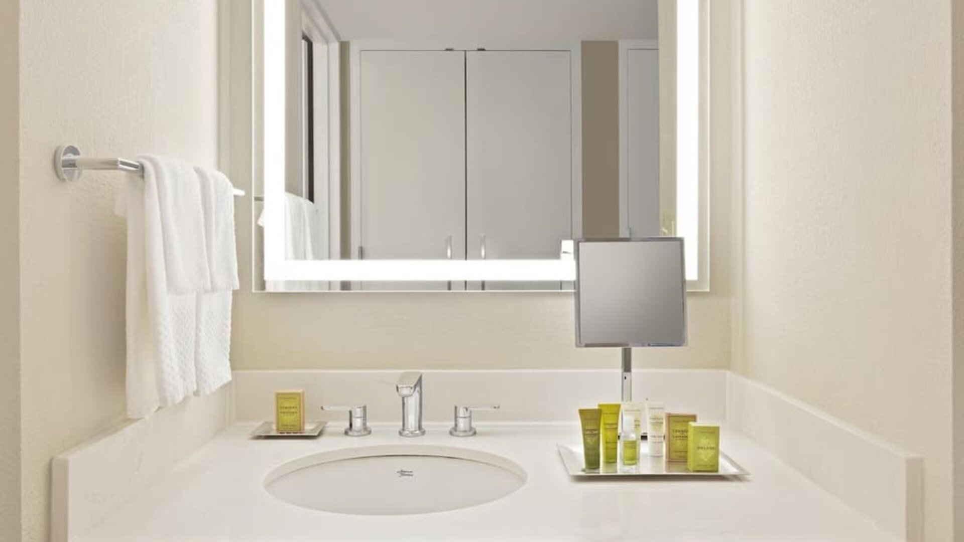 Bright hotel bathroom vanity with white countertop and sink, illuminated rectangular mirror, wall-mounted magnifying mirror, neatly arranged toiletries, and white towels.