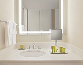 Bright hotel bathroom vanity with white countertop and sink, illuminated rectangular mirror, wall-mounted magnifying mirror, neatly arranged toiletries, and white towels.