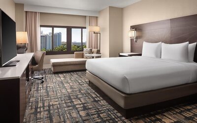 Contemporary hotel room with king bed in crisp white linens, large flat-screen TV, work desk and chair, chaise lounge, patterned carpet, and window overlooking nearby buildings and greenery.