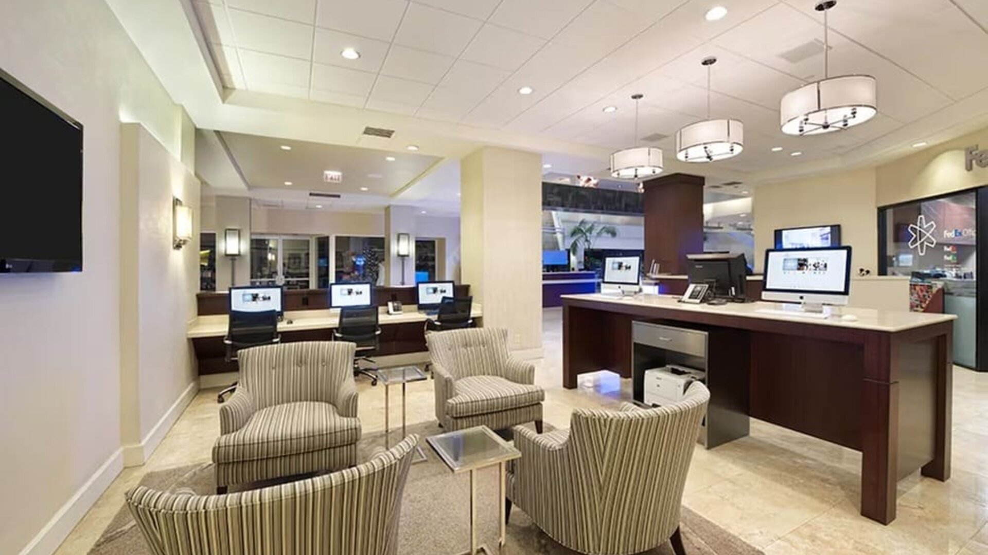 Spacious hotel business center with multiple desktop computers, a printer station, striped lounge chairs, small tables, and modern pendant lighting.