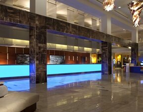 Contemporary hotel lobby with long illuminated blue reception counters, glossy tile flooring, dark architectural columns, modern hanging lights, and seating areas.