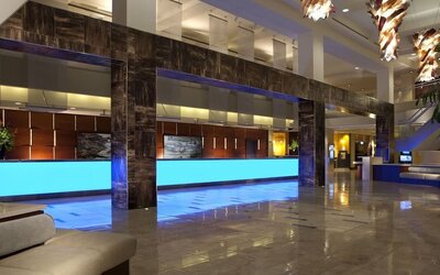 Contemporary hotel lobby with long illuminated blue reception counters, glossy tile flooring, dark architectural columns, modern hanging lights, and seating areas.
