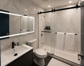 Bright modern bathroom with marble-look tile walls, illuminated mirror, dark vanity, toilet, and glass shower enclosure with black hardware and white towels.