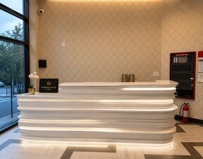 Fordham Hotel reception area with a sculpted white illuminated front desk, patterned beige walls, large street-facing windows, and hotel signage.
