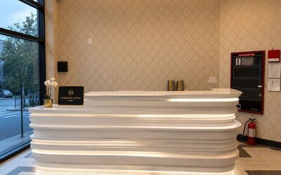 Fordham Hotel reception area with a sculpted white illuminated front desk, patterned beige walls, large street-facing windows, and hotel signage.
