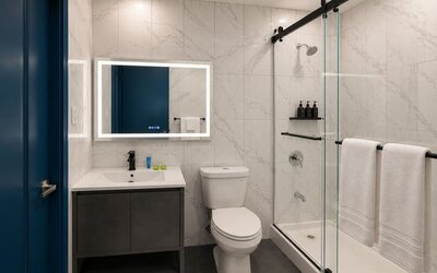 Modern hotel bathroom with marble-look wall tiles, illuminated mirror, white vanity, toilet, and glass-enclosed shower with black fixtures and white towels.