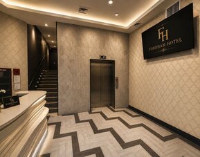 Fordham Hotel lobby with curved white reception desk, elevator, illuminated staircase, patterned tile flooring, decorative walls, and large hotel sign.