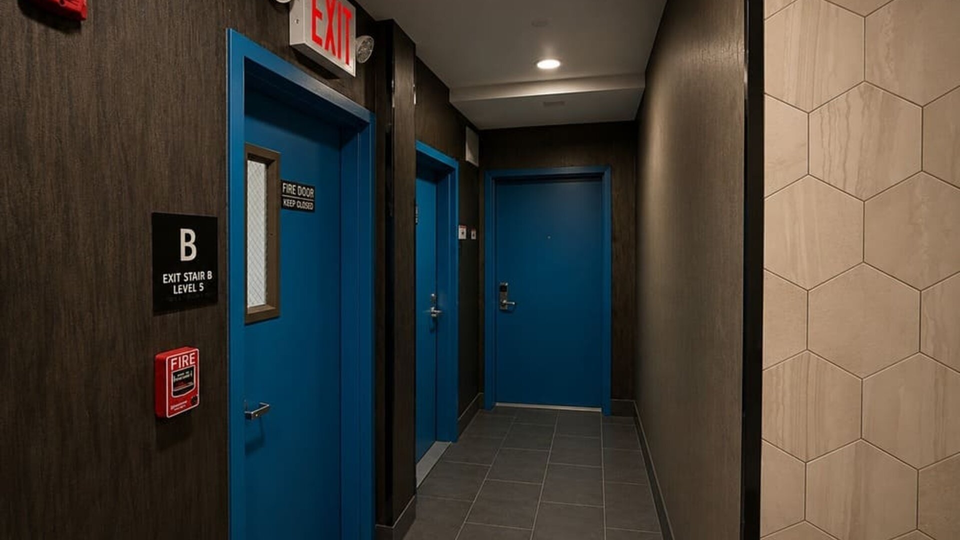 Dark contemporary hotel corridor with blue guest and exit doors, gray tiled flooring, recessed ceiling lights, and clearly marked fire and exit signage.