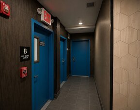 Dark contemporary hotel corridor with blue guest and exit doors, gray tiled flooring, recessed ceiling lights, and clearly marked fire and exit signage.